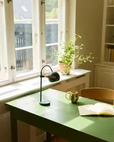 DC Portable Bordlampe Mørkegrøn fra Halo Design på et skrivebord ved et vindue